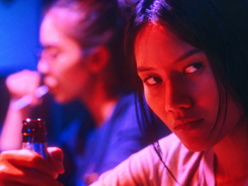 Copyright: 2026 eksystent Filmverleih Nahaufnahme einer jungen Frau in rot-blauem Neonlicht, die mit ernstem Blick zur Seite schaut, im Hintergrund eine Person mit einer Getränkeflasche.