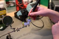 Eine Person lötet ein kleines, cartoonhaftes Einhorn-Schmuckstück aus Metall, das in einem Hilfsarm eingespannt ist.