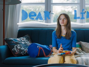 Copyright: Veletlenuel Film; Beta Film
Eine junge Person sitzt nachdenklich mit Notizbuch auf einem blauen Sofa.
