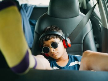 Copyright: Aditya Varma Kind mit roter Kopfhörern und runden Sonnenbrille lehnt entspannt auf dem Rücksitz eines Autos, während gelb-lila Socken in den Vordergrund ragen.