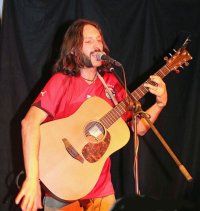 Guy Palumbo spielt leidenschaftlich Akustikgitarre und singt in ein Mikrofon auf einem Ständer.