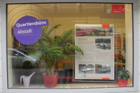 Schaufenster des Quartiersbüros Altstadt Nürnberg mit lila Logo, einer Infotafel zum Cramer-Klett-Park und Zimmerpflanzen.LONGDESC: Das Bild zeigt ein Schaufenster eines Büros. Linke Seite: Auf der Glasscheibe ist ein lila Kreis mit konzentrischen lila Ringen aufgebracht. Im Kreis steht in weißer Schrift: „Quartiersbüro, Altstadt". Oben rechts auf der Scheibe: rotes Logo mit Stadtsilhouette und Schriftzug „NÜRNBERG". Mitte/rechts: Im Schaufenster steht eine transparente Halterung mit einer Infotafel. Die Tafel zeigt oben dasselbe rote Nürnberg-Logo sowie die Aufschrift „Quartiersbüro, Altstadt". Darunter eine Überschrift (teilweise lesbar): „Projekt der Stadterneuerung, Cramer-Klett-Park". Es folgen mehrere Textabsätze (zu klein/unscharf für vollständige Transkription) sowie zwei historische Schwarz-Weiß-Fotos und zwei Farbfotos des Parks. Unten auf der Tafel sind kleine Logos erkennbar (unleserlich, zu klein). Im Schaufenster steht außerdem eine große Zimmerpflanze (Palme) in einem roten Topf sowie eine kleinere Pflanze rechts. Links daneben ist eine weiße Gießkanne zu sehen.