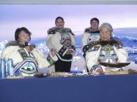Vier Frauen in traditionell bestickter Kleidung mit Fellbesatz stehen und sitzen an einem Tisch mit kunsthandwerklichen Objekten, im Hintergrund ein Bildschirm mit Eislandschaft.