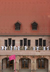Mehrere Personen in weißen und goldenen Anzügen stehen auf dem Balkon über dem Haupteingang des Künstlerhauses und winken