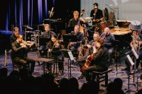 Kammerensemble aus rund zwölf Personen musiziert auf einer Bühne mit Streichern, Bläsern und Schlagwerk vor Publikum.