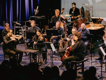 Kammerensemble aus rund zwölf Personen musiziert auf einer Bühne mit Streichern, Bläsern und Schlagwerk vor Publikum.