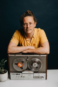 Person mit Brille und hochgesteckten Locken lehnt die Arme auf einem vintage Philips-Tonbandgerät (4404 Stereo, 4-Track) und blickt ruhig in die Kamera.
