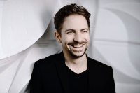 Lachende Person mit braunen Haaren, grünen Augen und Bart in schwarzem Outfit vor weißem Hintergrund.