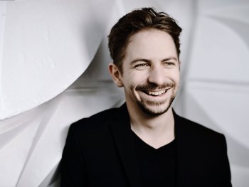 Lachende Person mit braunen Haaren, grünen Augen und Bart in schwarzem Outfit vor weißem Hintergrund.