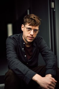 Eine junge Person mit Brille und braunem, leicht zerzaustem Haar sitzt in nachdenklicher Pose vor einem dunklen Hintergrund.