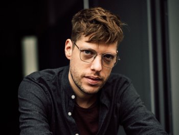 Eine junge Person mit Brille und braunem, leicht zerzaustem Haar sitzt in nachdenklicher Pose vor einem dunklen Hintergrund.