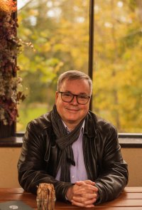 Lächelnde Person mit Brille und grauem Schal sitzt in einem Café an einem Holztisch vor einem Fenster mit herbstlich gefärbten Bäumen.
