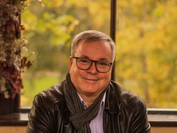 Lächelnde Person mit Brille und grauem Schal sitzt in einem Café an einem Holztisch vor einem Fenster mit herbstlich gefärbten Bäumen.