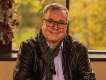 Lächelnde Person mit Brille und grauem Schal sitzt in einem Café an einem Holztisch vor einem Fenster mit herbstlich gefärbten Bäumen.