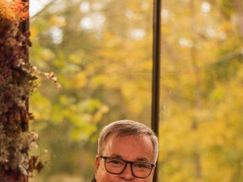 Foto: Stella Reiss
Lächelnde Person mit Brille und grauem Schal sitzt in einem Café an einem Holztisch vor einem Fenster mit herbstlich gefärbten Bäumen.