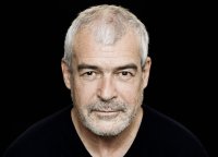 Porträtfoto einer älteren Person mit kurzen grauen Haaren und Dreitagebart, die ernst und direkt in die Kamera blickt.