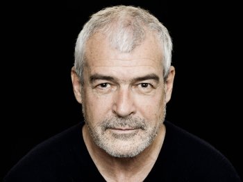 Porträtfoto einer älteren Person mit kurzen grauen Haaren und Dreitagebart, die ernst und direkt in die Kamera blickt.