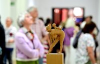 Holzfigur einer sich vorbeugenden Person auf einem Sockel im Vordergrund einer belebten Kunstausstellung mit unscharfen Besucherinnen und Besuchern im Hintergrund.