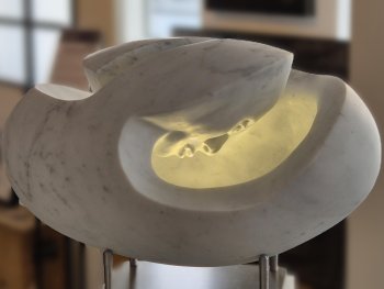 Eiförmige Marmorskulptur mit innen leuchtendem, warmgelbem Licht, das auf ein Gesicht im inneren scheint.