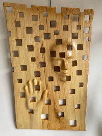 Holzskulptur mit aus dem Holz herausgearbeiteten Gesicht und Hand einer Person, umgeben von regelmäßig angeordneten quadratischen Aussparungen.