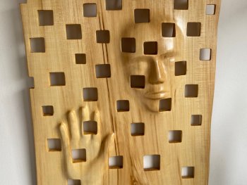 Holzskulptur mit aus dem Holz herausgearbeiteten Gesicht und Hand einer Person, umgeben von regelmäßig angeordneten quadratischen Aussparungen.