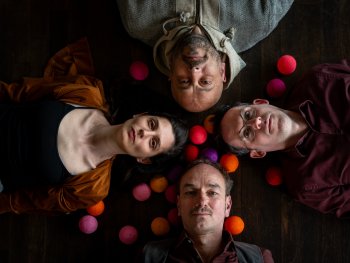 Vier Personen liegen mit den Köpfen zur Mitte auf einem Holzboden, umgeben von bunten Wollbällen in Rosa, Orange, Rot und Lila.