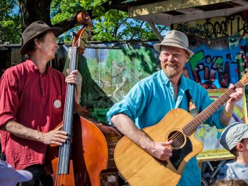 Zwei Personen mit Hüten spielen lächelnd bei einem Outdoor-Konzert Akustikgitarre und Kontrabass vor einer bemalten Wand.