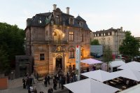 Außenansicht der Kunstvilla Nürnberg bei Dämmerung mit beleuchtetem Gründerzeitbau und Personen sowie weißen Marktschirmen auf dem Vorplatz.