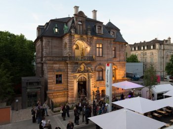 Foto: Kunstvilla/Annette Kradisch
Außenansicht der Kunstvilla Nürnberg bei Dämmerung mit beleuchtetem Gründerzeitbau und Personen sowie weißen Marktschirmen auf dem Vorplatz.
