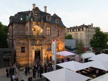 Außenansicht der Kunstvilla Nürnberg bei Dämmerung mit beleuchtetem Gründerzeitbau und Personen sowie weißen Marktschirmen auf dem Vorplatz.