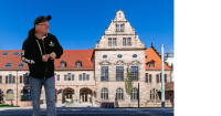 Eine Person in schwarzem Kapuzenpullover mit Totenkopf-Logo steht vor einem historischen Sandsteingebäude mit rotem Ziegeldach und gestuftem Treppengiebel.