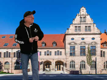 Eine Person in schwarzem Kapuzenpullover mit Totenkopf-Logo steht vor einem historischen Sandsteingebäude mit rotem Ziegeldach und gestuftem Treppengiebel.