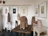 Skulpturenausstellung mit mehreren abstrakten Figuren aus Ton auf Tischen und Podesten in einem weißen Raum mit gerahmten Zeichnungen an den Wänden