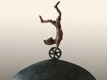 Bronzeskulptur einer akrobatisch kopfstehenden Person auf einem Einrad, das auf einem halbkugelförmigen grünen Serpentinsockel balanciert.
