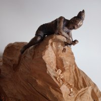 Bronzefigur einer Person, die auf einer hölzernen Skulptur rittlinks sitzt und diese bearbeitet.