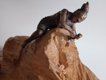 Bronzefigur einer Person, die auf einer hölzernen Skulptur rittlinks sitzt und diese bearbeitet.