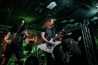 Drei Personen mit langen Haaren spielen E-Gitarren auf einer grün beleuchteten Bühne in einem kleinen Club; eine Person singt ins Mikrofon.