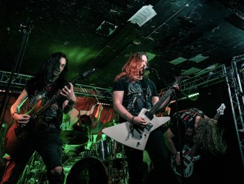 Drei Personen mit langen Haaren spielen E-Gitarren auf einer grün beleuchteten Bühne in einem kleinen Club; eine Person singt ins Mikrofon.