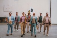 Sieben Personen in eleganter Freizeitkleidung gehen mit Blasinstrumenten – darunter Posaune, Saxofon, Tuba und Trompete – auf die Kamera zu