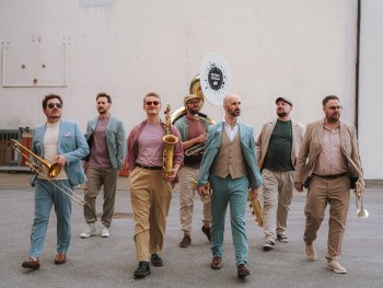 Sieben Personen in eleganter Freizeitkleidung gehen mit Blasinstrumenten – darunter Posaune, Saxofon, Tuba und Trompete – auf die Kamera zu