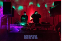 Zwei DJs legen an Turntables auf, während rotes und grünes Licht die Bühne beleuchtet, darunter der Schriftzug „Disko Disko“.