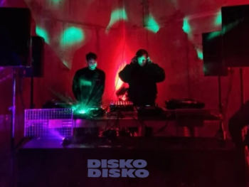 Zwei DJs legen an Turntables auf, während rotes und grünes Licht die Bühne beleuchtet, darunter der Schriftzug „Disko Disko“.