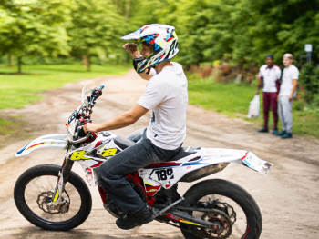Copyright: Grandfilm; Vajda film Person mit Motocross-Helm sitzt auf einem Motorrad mit Startnummer 189 auf einem Feldweg im Grünen, im Hintergrund stehen weitere Personen.