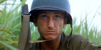 Ein Soldat mit Stahlhelm und Gewehr kauert schwitzend im hohen Gras und blickt angespannt in die Ferne.
