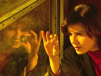 Copyright: Studiocanal Person mit dunklen Haaren betrachtet an einem Fenster eine kleine Glaskugel zwischen den Fingern in warmem Licht.