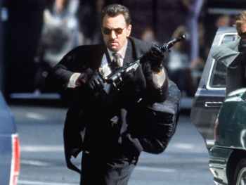 Copyright: Warner Bros
Zwei bewaffnete Männer in Anzügen mit Sonnenbrillen und Maschinengewehren bei einer Schießerei auf einer Straße neben einem Fahrzeug.