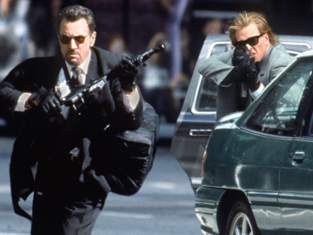 Copyright: Warner Bros Zwei bewaffnete Männer in Anzügen mit Sonnenbrillen und Maschinengewehren bei einer Schießerei auf einer Straße neben einem Fahrzeug.