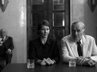 Schwarz-weißes Filmszenenfoto zeigt eine Frau und einen Mann mit ernster Mimik nebeneinander an einem Tisch mit Trinkgläsern sitzend, im Hintergrund eine weitere Person mit Amtskette.