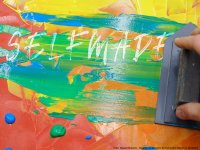 Hand mit Spachtel verteilt bunte Farbe auf einer roten Unterlage mit dem weißen Schriftzug 