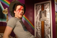 Eine Person mit rot-blauem Blitz-Gesichtsschminke im Stil von David Bowie sitzt in einem lila gestrichenen Zimmer mit Regenbogen-Wandmalerei und einer mit Alufolie verzierten Tür.
